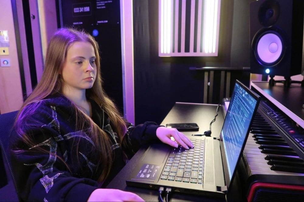 A young hip hop producer working in a recording studio at a laptop beside a keyboard, lit by purple ambient light.