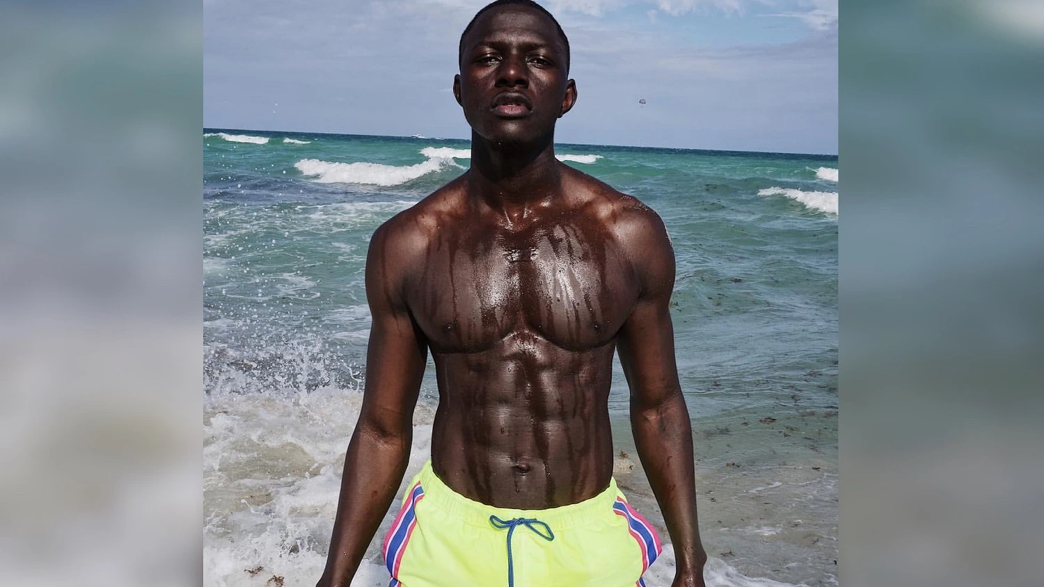 Mohamet Mbaye standing shirtless on a beach with ocean waves behind him