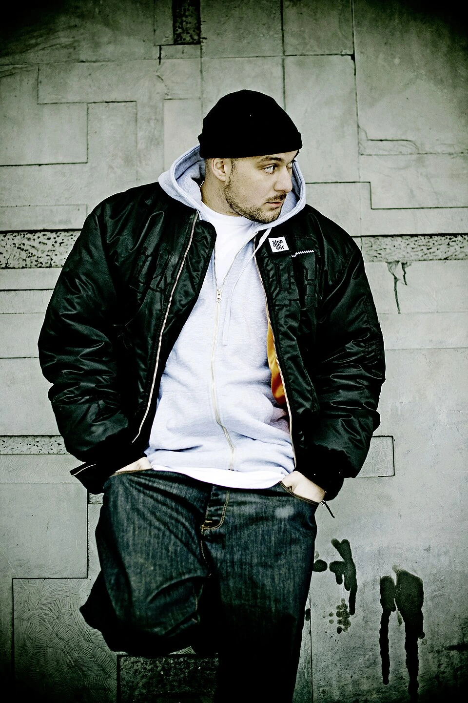 Kool Savas posing in a black jacket and beanie against a concrete wall
