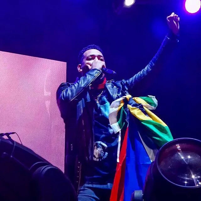 AKA performing on stage at Oppikoppi 2015, holding a microphone with one arm raised.