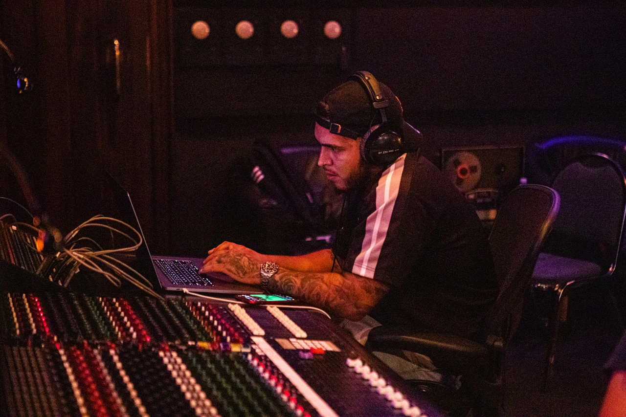 Vybe Beatz seated at a mixing console in a recording studio, wearing headphones and working on a laptop