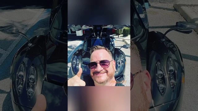 A man taking a selfie in front of a sports car, giving a thumbs-up.