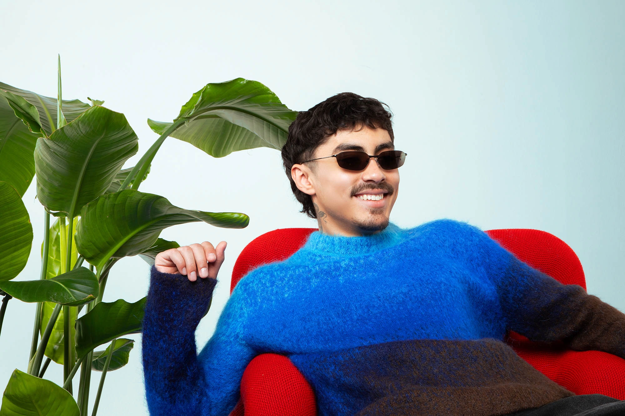 Funk Tribu smiling in a studio portrait, wearing sunglasses and a blue sweater beside large green leaves.