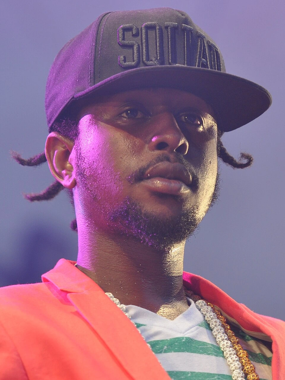 Popcaan performing on stage, wearing a cap and colorful jacket under concert lighting.