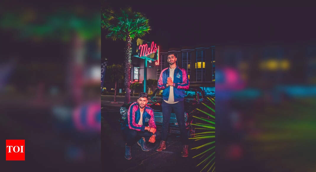 Two men in matching blue-and-pink jackets posing at night beside a car under neon signage, representing the twin DJ/prod